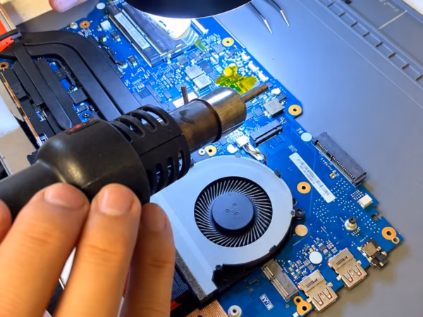 Technician using a hot-air rework wand on a laptop motherboard beneath a digital inspection microscope.